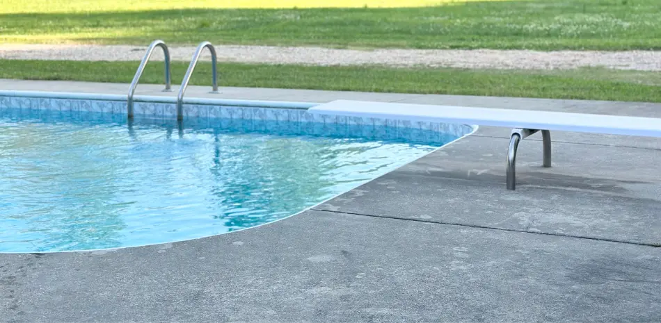 A calm outdoor pool with clear blue water, a metal ladder, and a white diving board, sits adjacent to a concrete poolside.